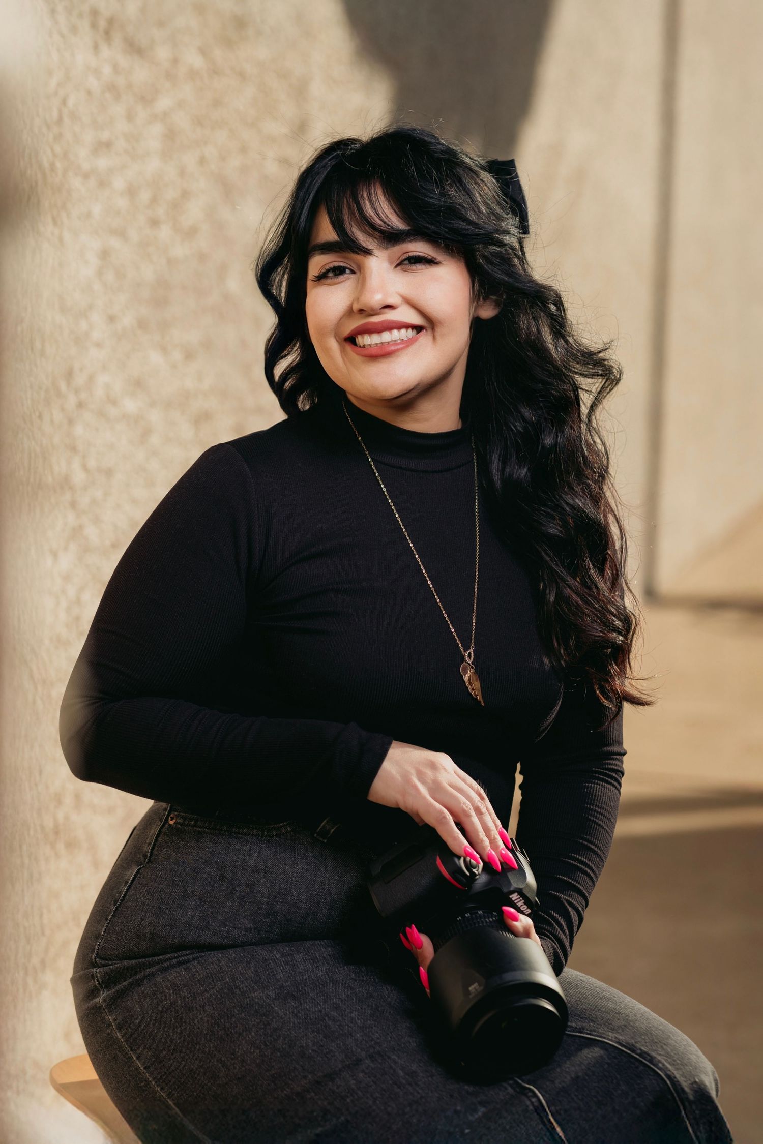 Portrait of a photographer in a black turtleneck holding a camera and smiling while sitting against a textured wall.