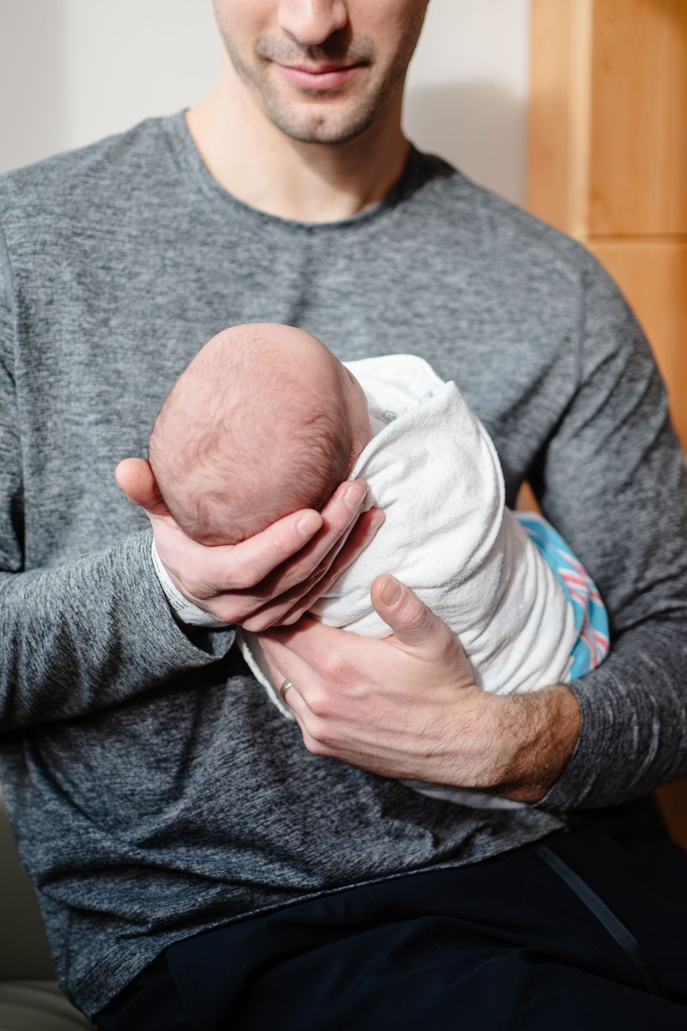 A parent in a gray sweater tenderly holds a newborn baby wrapped in a white blanket.