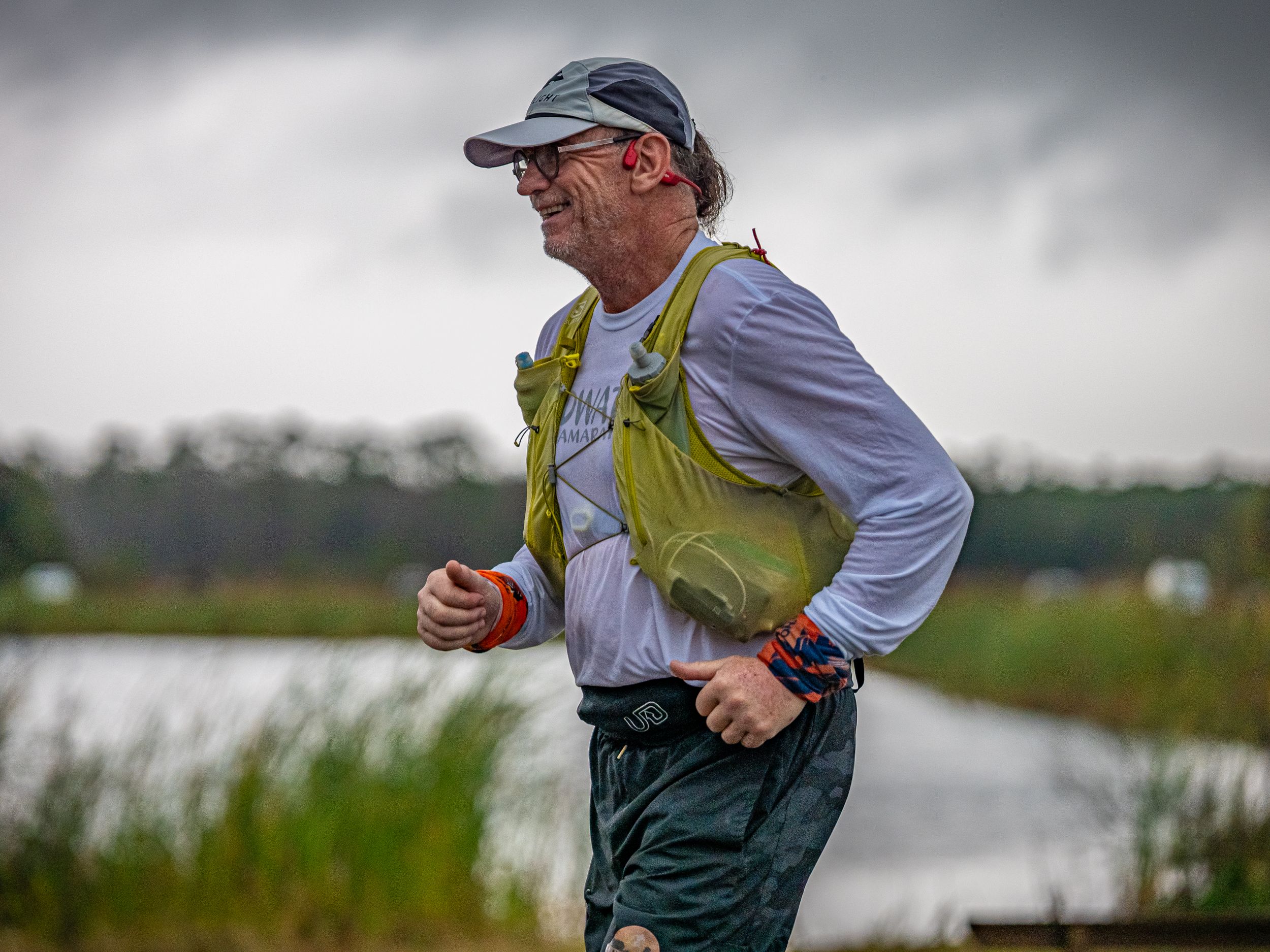 Runner in yellow safety vest and baseball cap moving along waterside trail during overcast conditions.