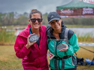 Two athletes display their finisher medals while smiling in outdoor running gear.