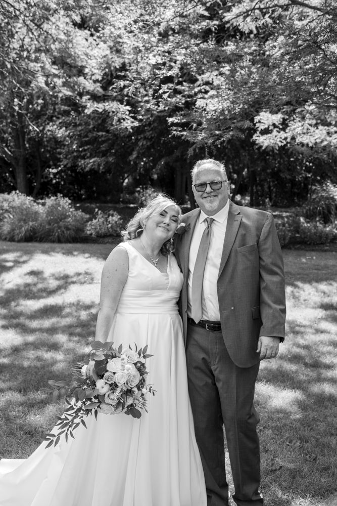 Black and white outdoor wedding portrait of couple on lawn with leafy trees in background.