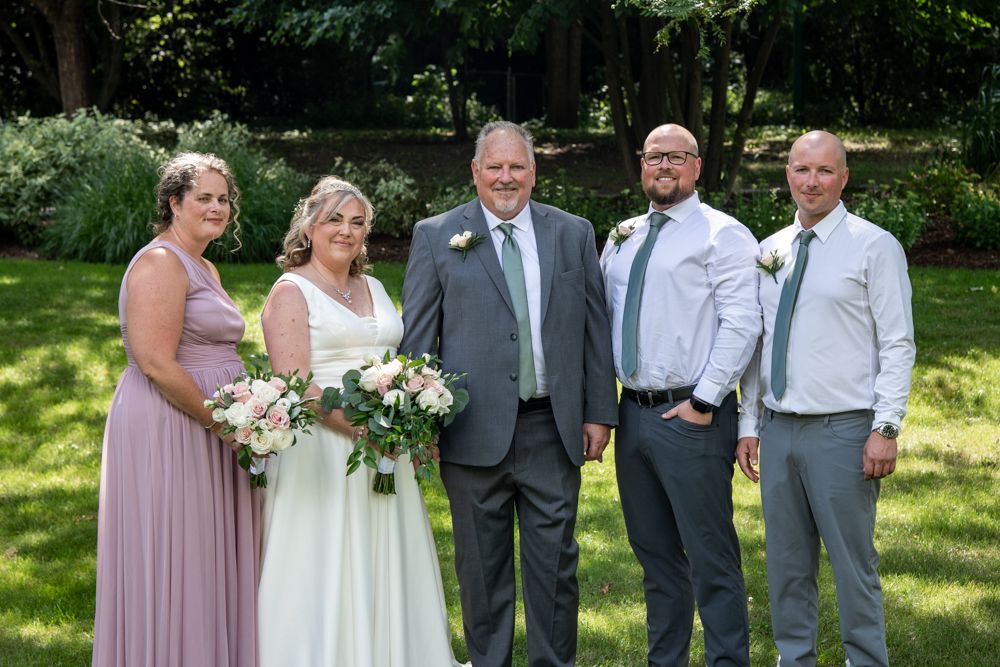 Wedding party in formal attire poses outdoors with floral bouquets in a sunny park setting.
