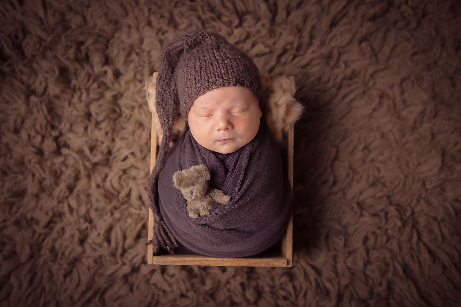 Sleeping newborn wrapped in purple swaddle rests in wooden prop bed on textured brown backdrop.