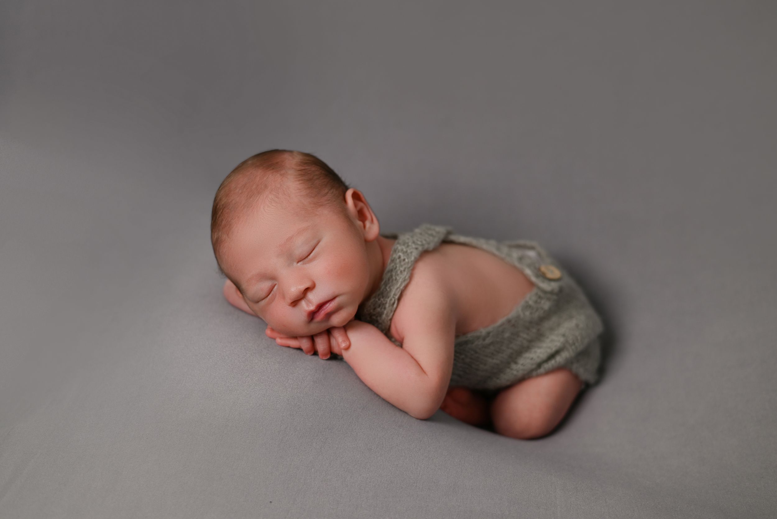 Newborn baby sleeping peacefully in grey outfit against neutral backdrop during professional photo session.