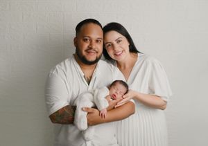 Happy couple in white outfits holding their sleeping newborn against light background.