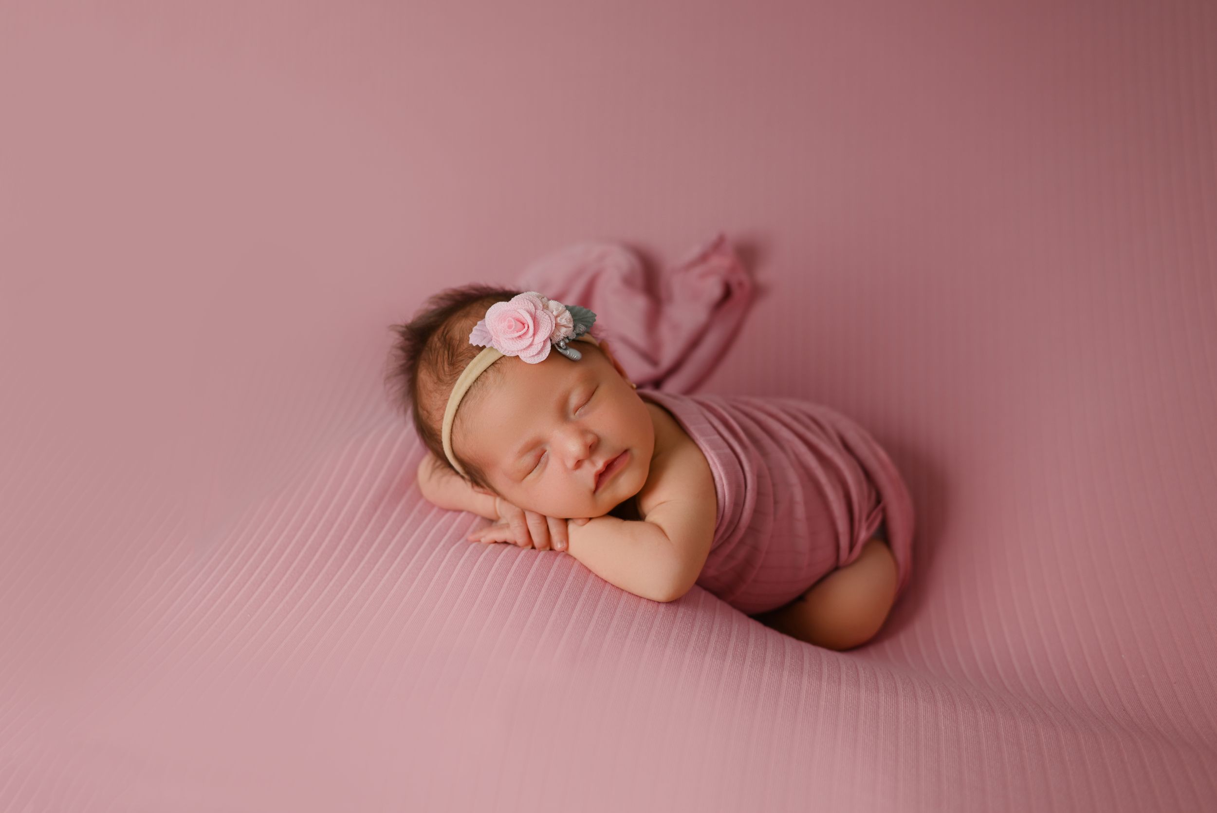 Sleeping newborn in pink wrap with delicate headband on pink textured backdrop.