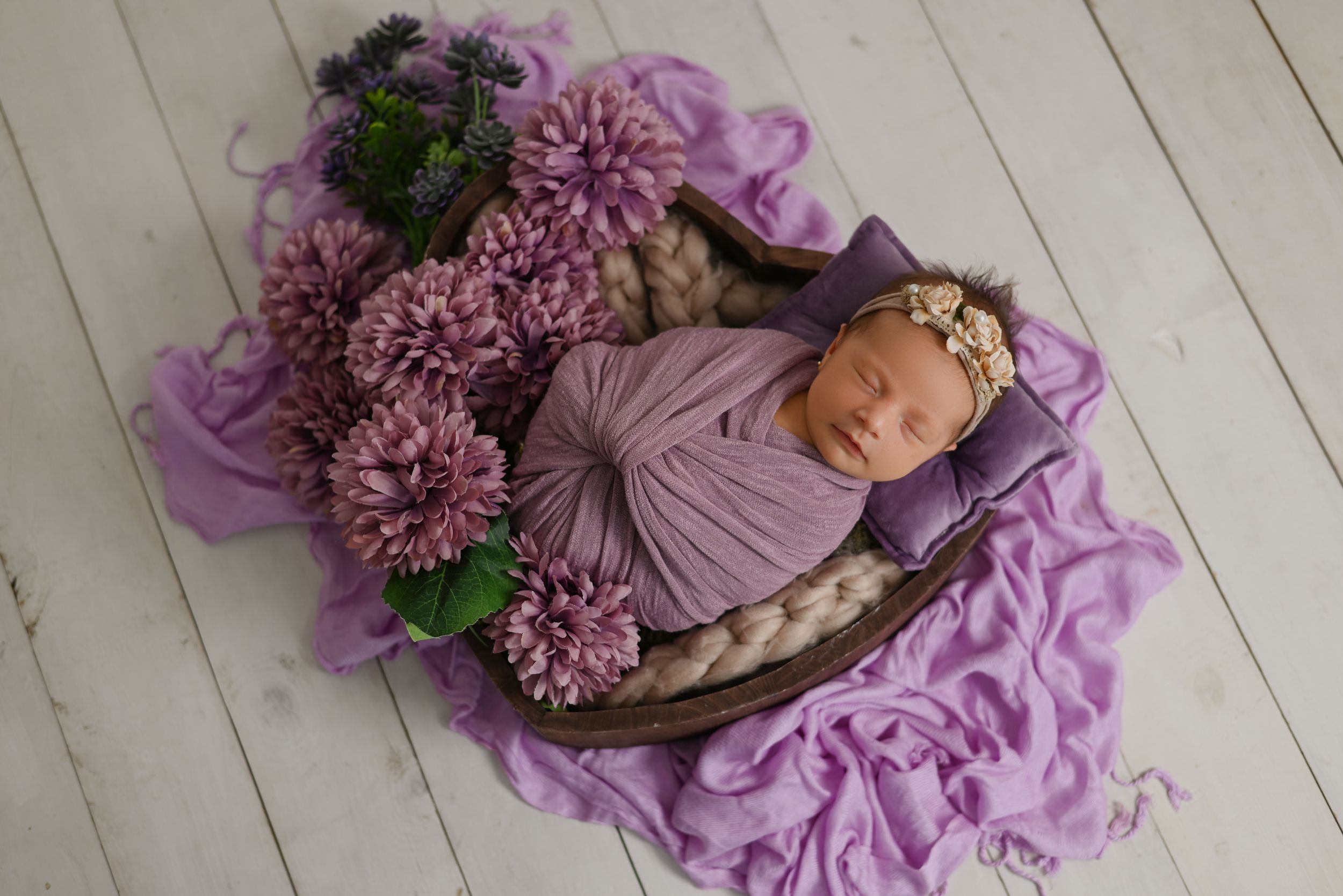 Newborn portrait with purple flowers and fabric in rustic wooden basket setup.