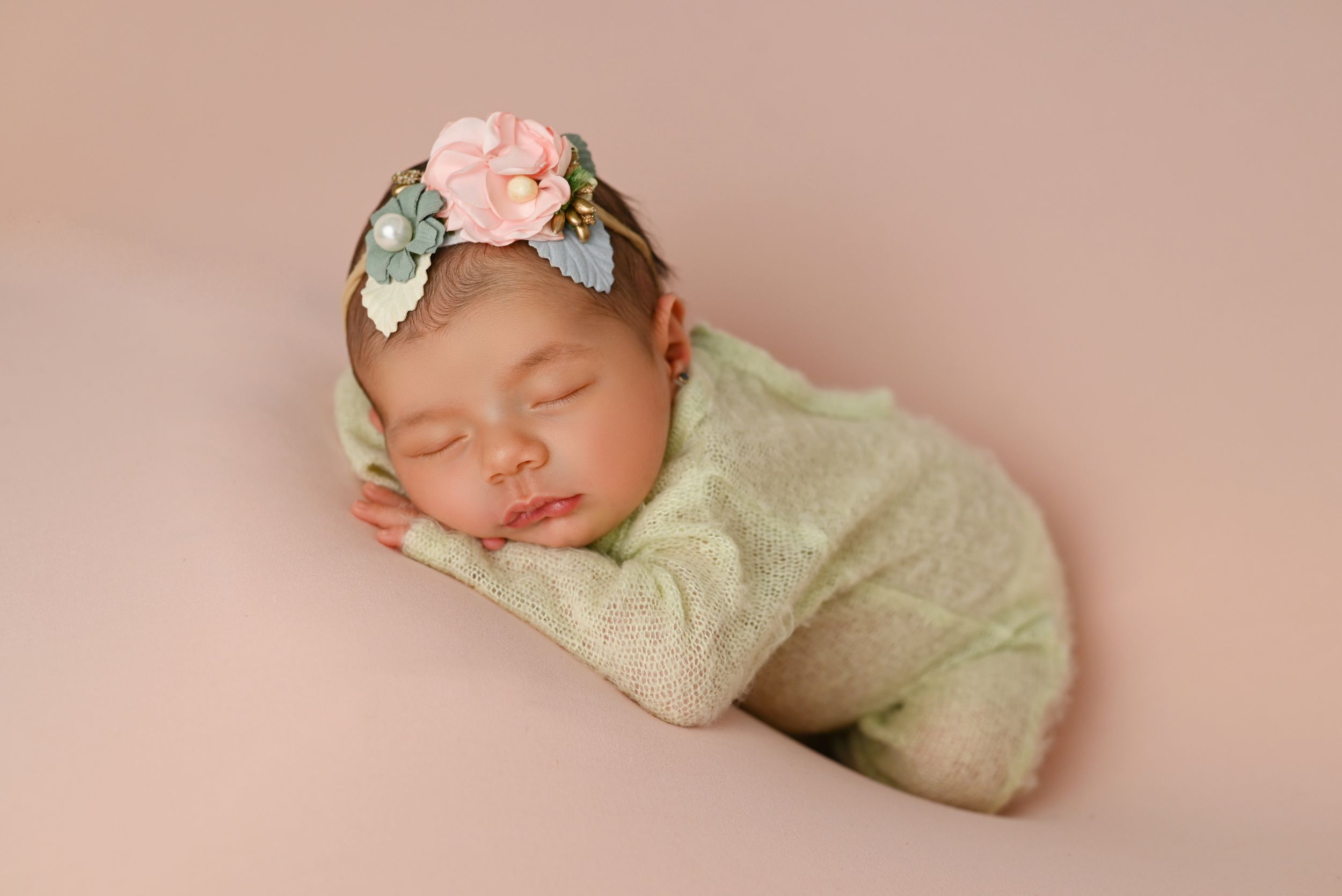 Sleeping newborn in mint outfit with floral headband on pink backdrop.