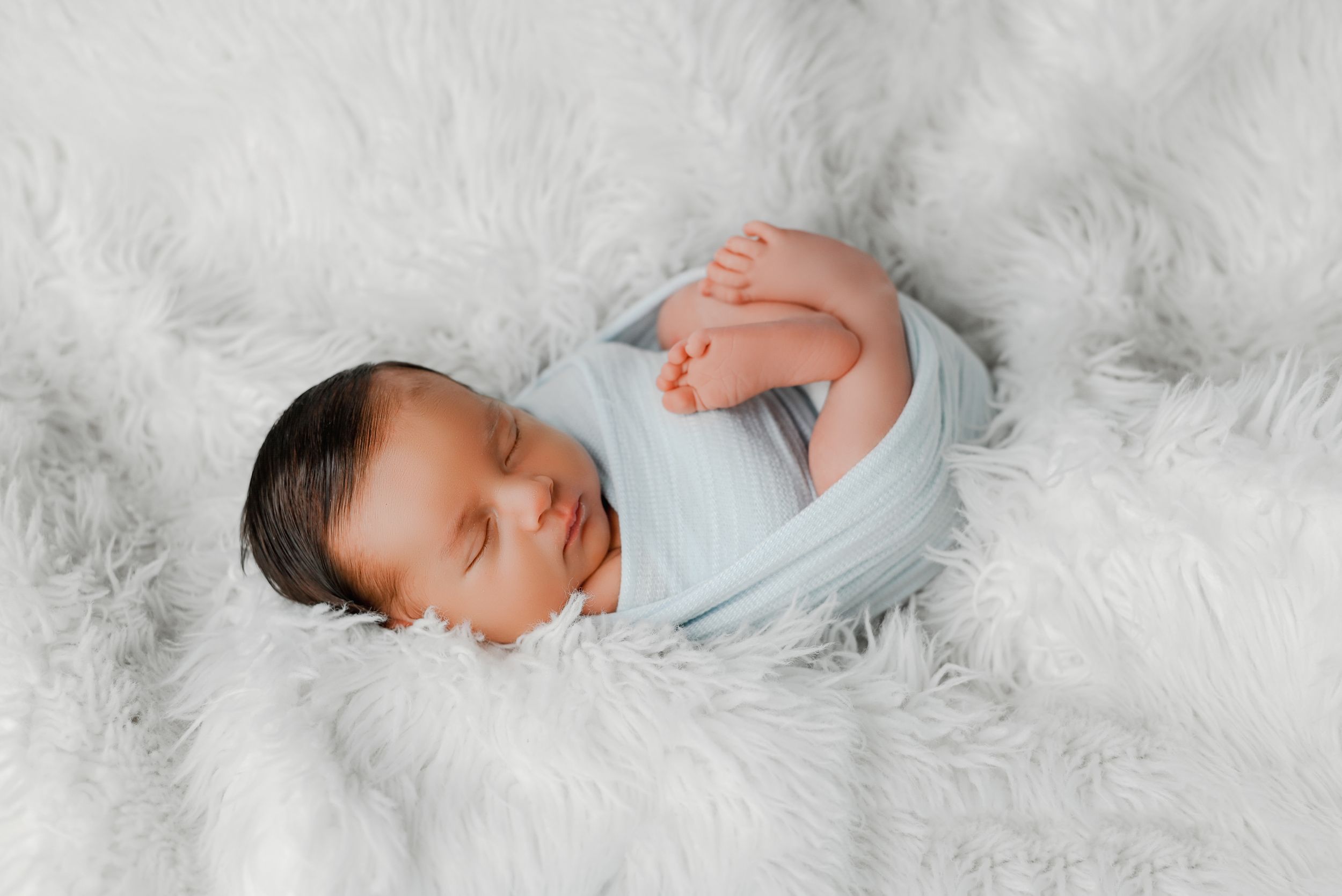 Peaceful newborn swaddled in light blue wrap on white furry background.