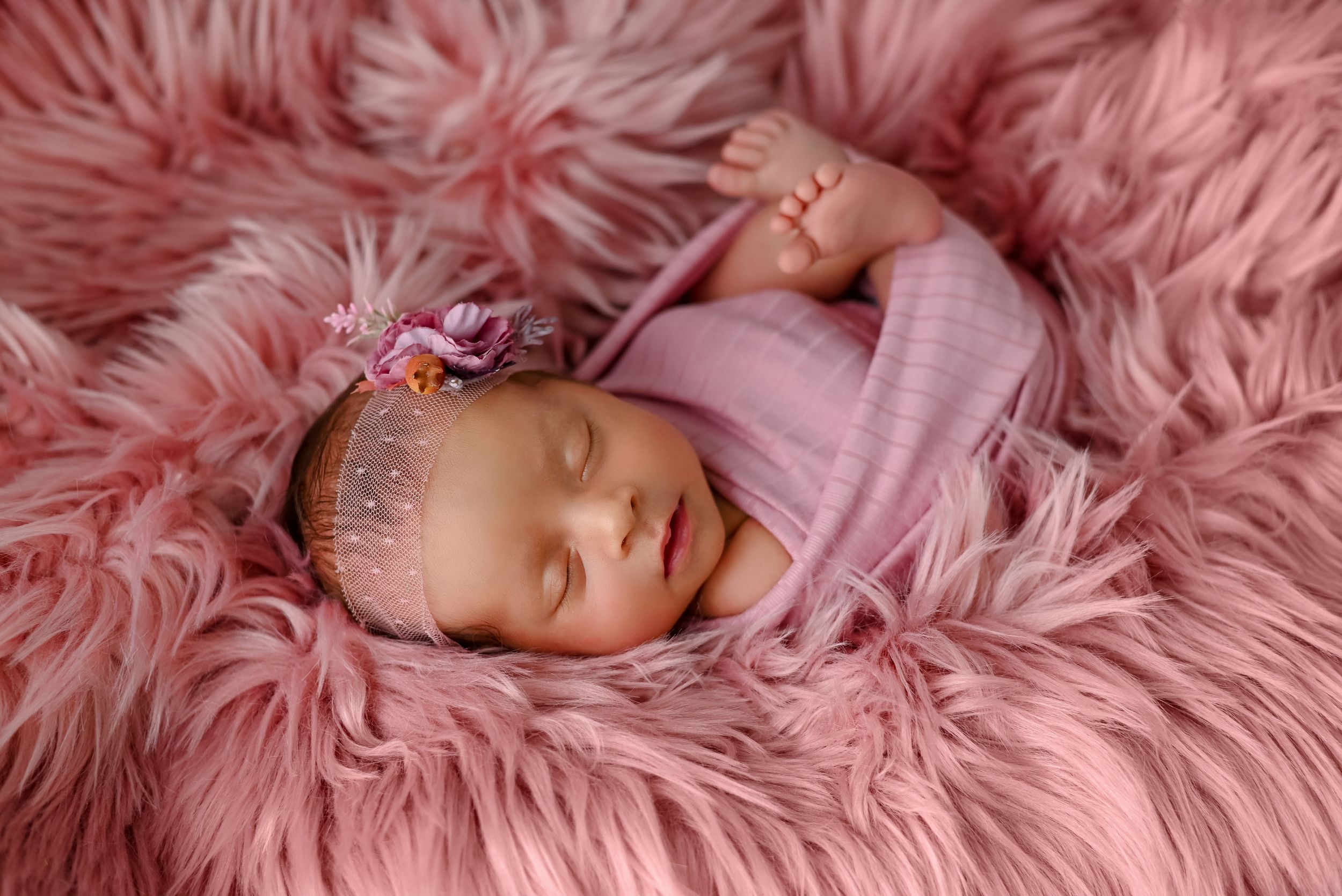 Sleeping baby in pink wrap on pink furry textile during newborn photo session.