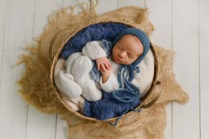 Sleeping newborn in white outfit nestled in blue fabric inside wooden bowl.