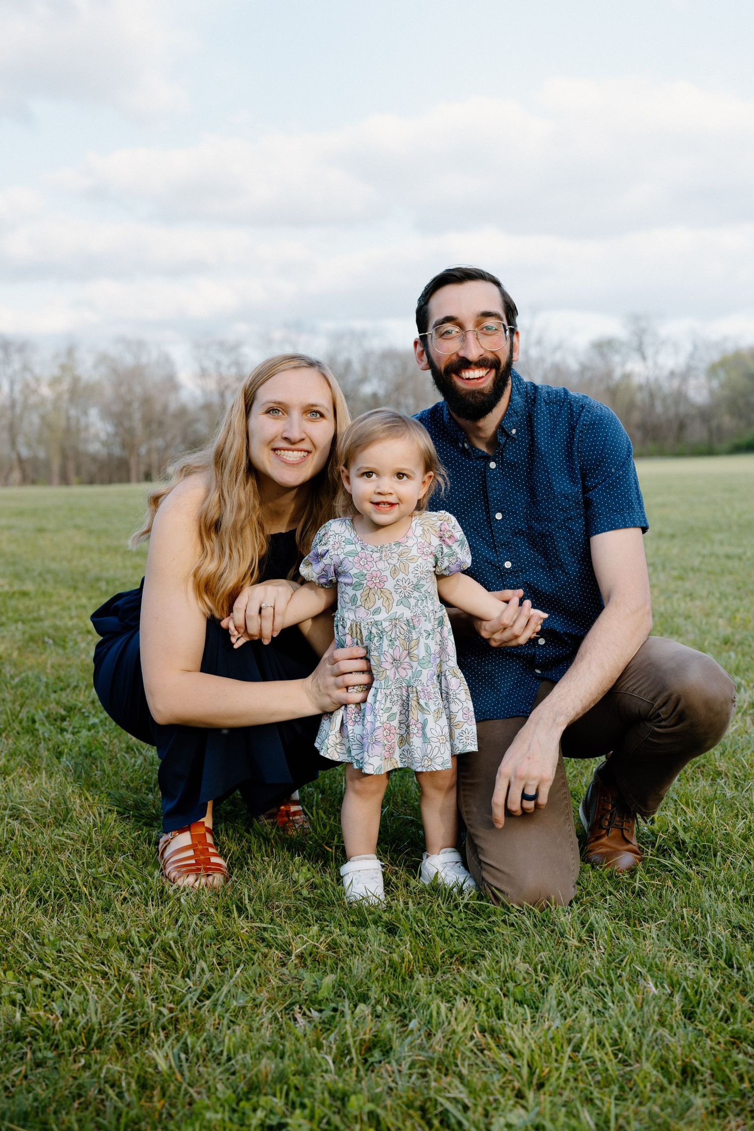 Family of three poses together on grass with trees in background.