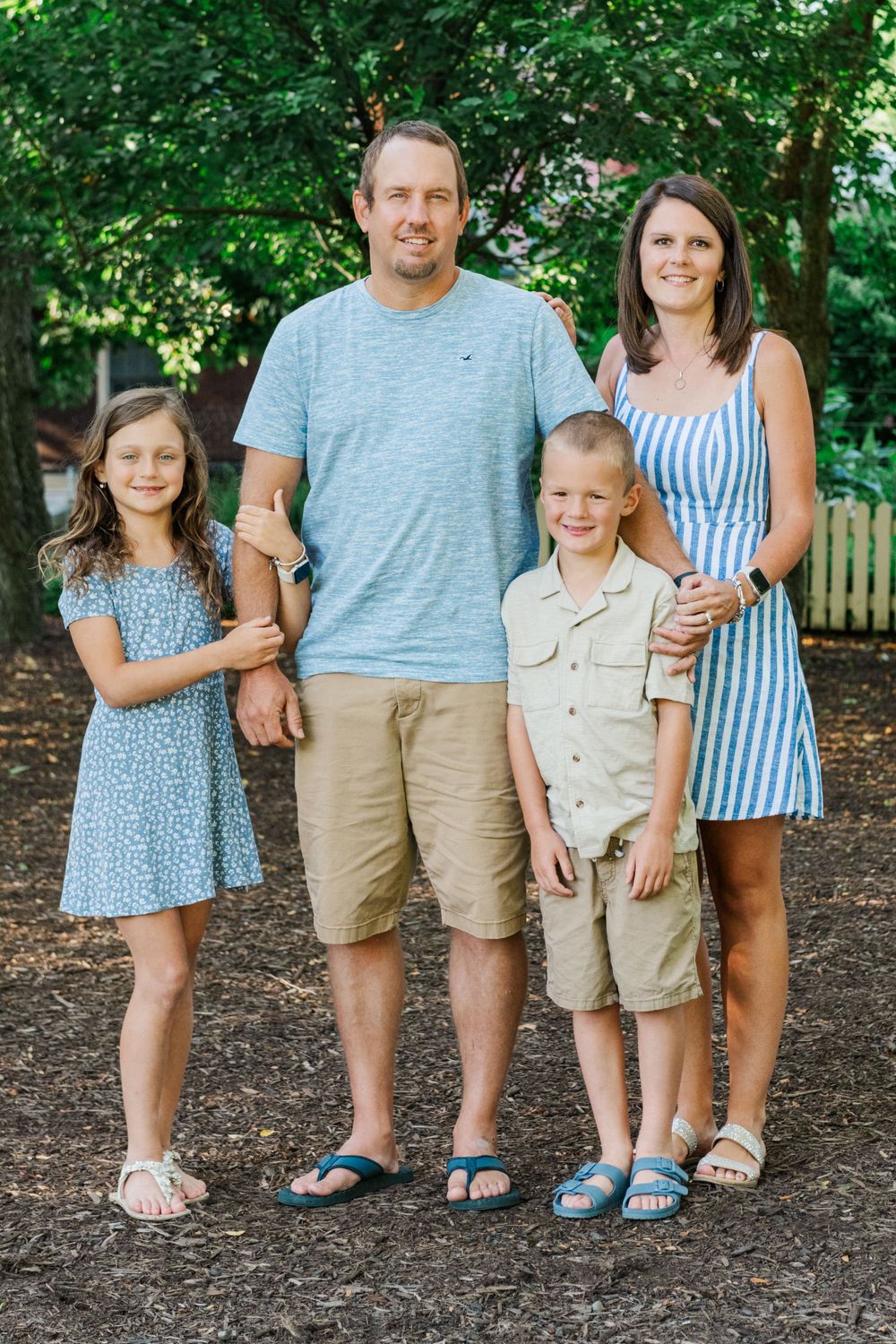 A family of four wearing coordinated blue and white outfits poses together outdoors with trees in the background.