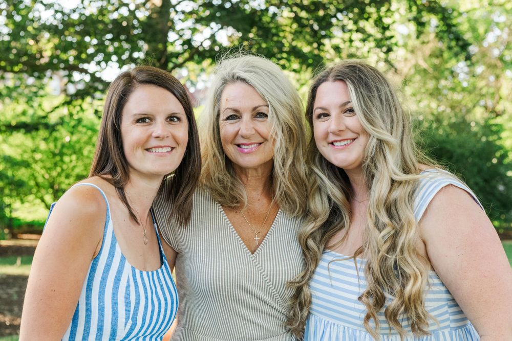 Three women wearing light colored tops smile together while posing outdoors in a garden setting.