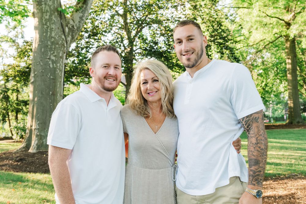 Three people stand together smiling outdoors in a park setting with green trees in the background.