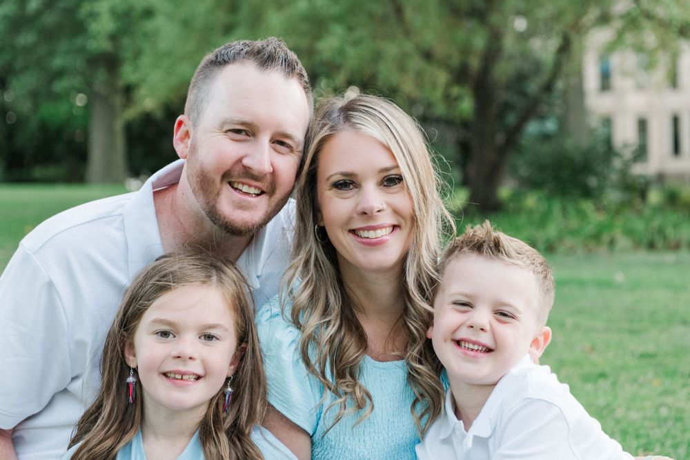 A happy family of four poses together outdoors in a sunny park with green trees in the background.