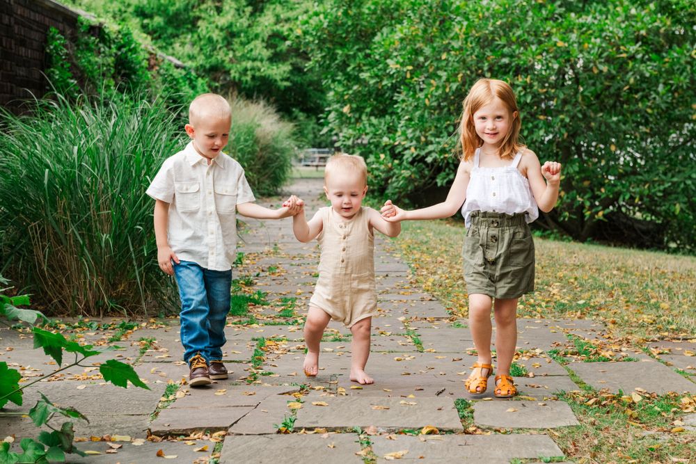 Three children holding hands and walking together on a stone path through a lush garden setting.