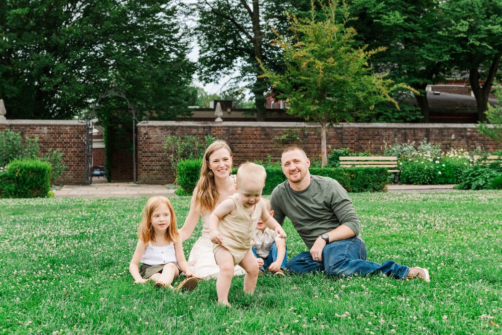 A family sits together on a green lawn in front of a brick wall and garden setting during a summer day.