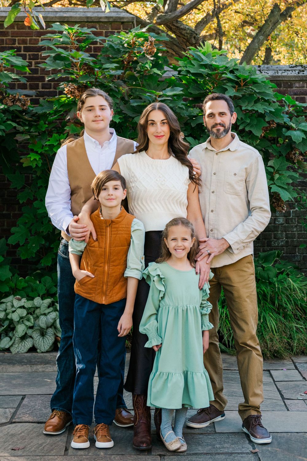A family of five poses together outdoors against a brick wall and green foliage on an autumn day.
