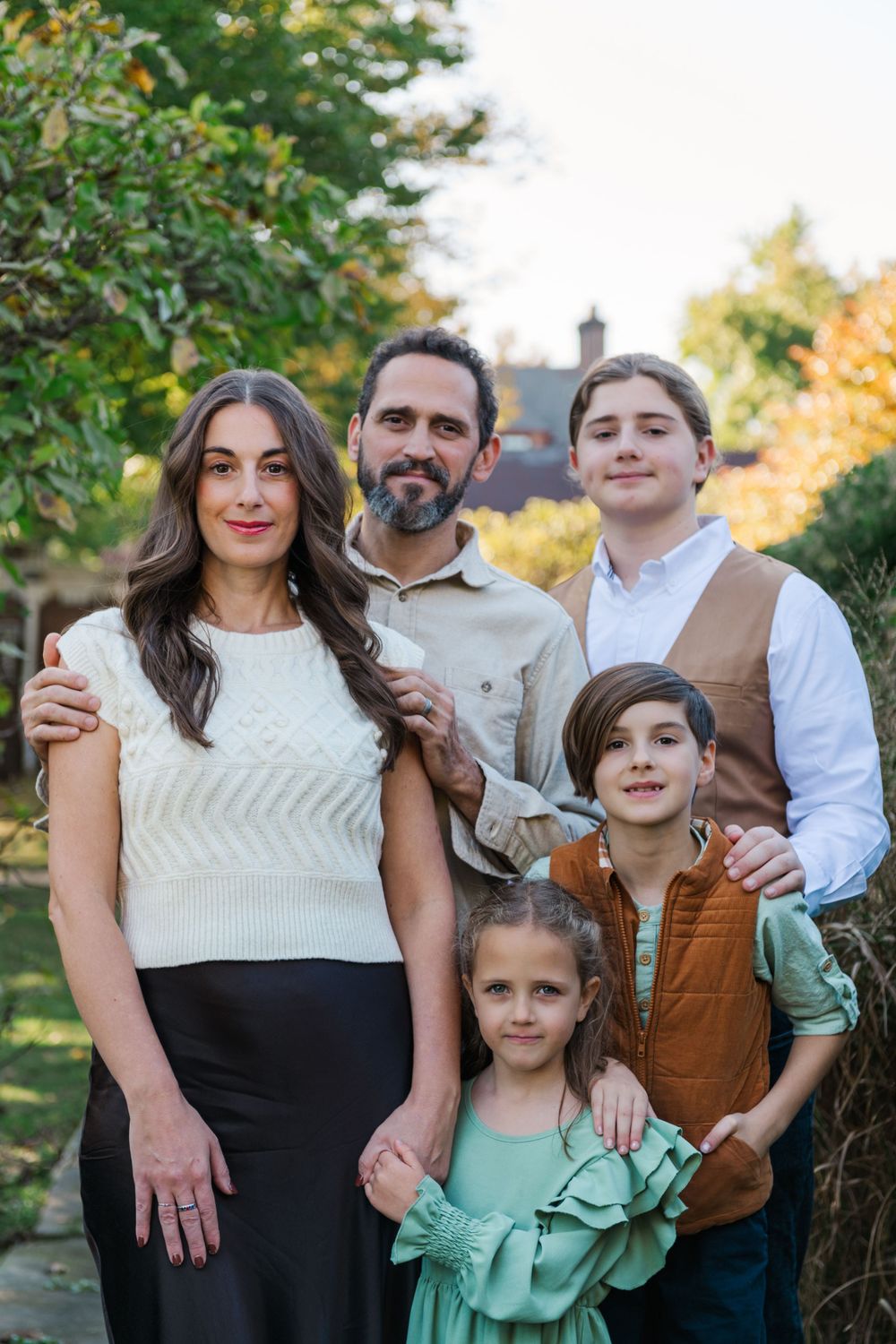 A family in coordinated autumn outfits stands together outdoors with fall foliage in the background.