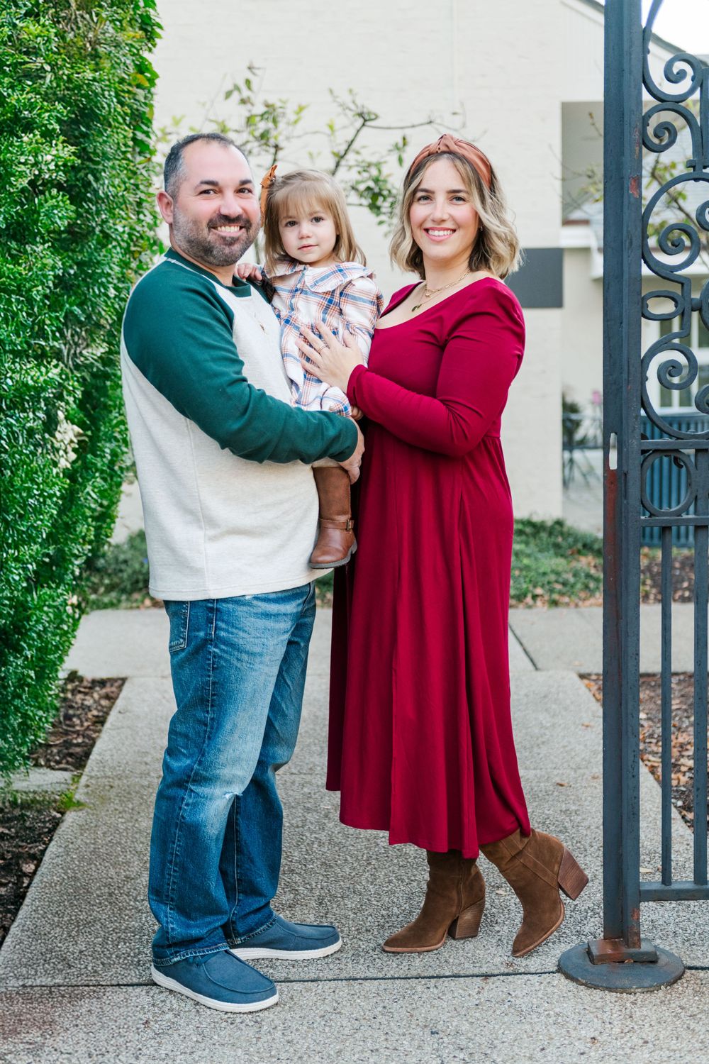 A family stands together beside a decorative iron gate, with the parent in green and white next to one in a red dress.