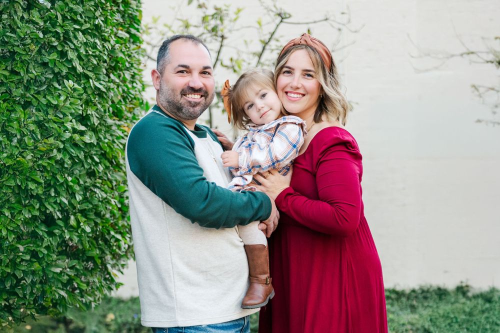 A cheerful family of three poses together outdoors against a white wall with green ivy.