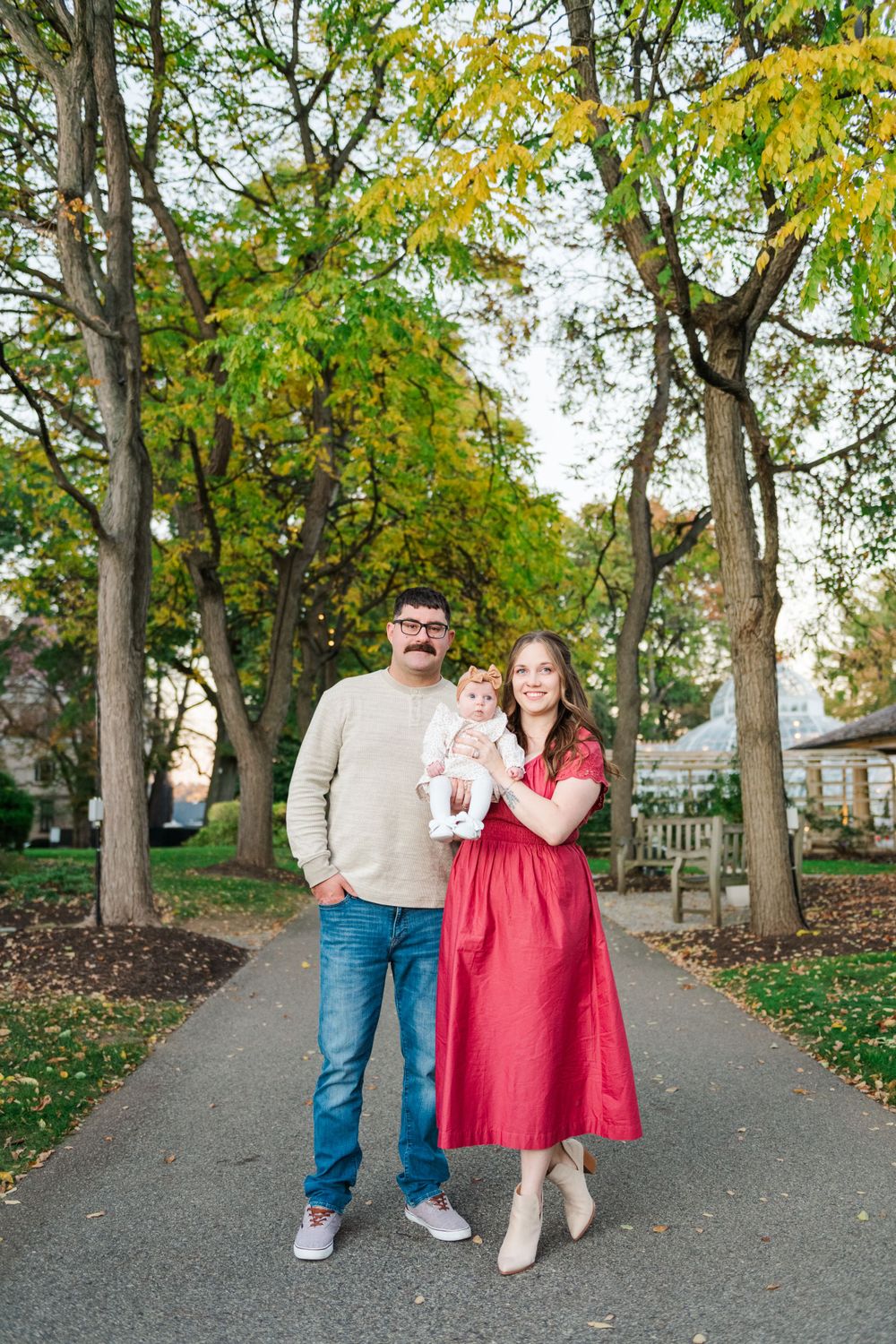 A couple stands together on a tree-lined autumn path with yellow leaves overhead.