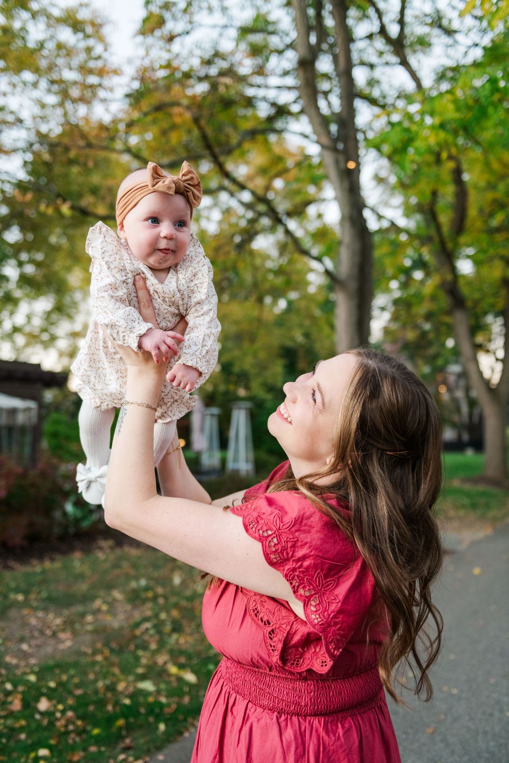 A baby in a lace outfit and bow headband is lifted high by someone wearing a rose-colored dress in an autumn setting.