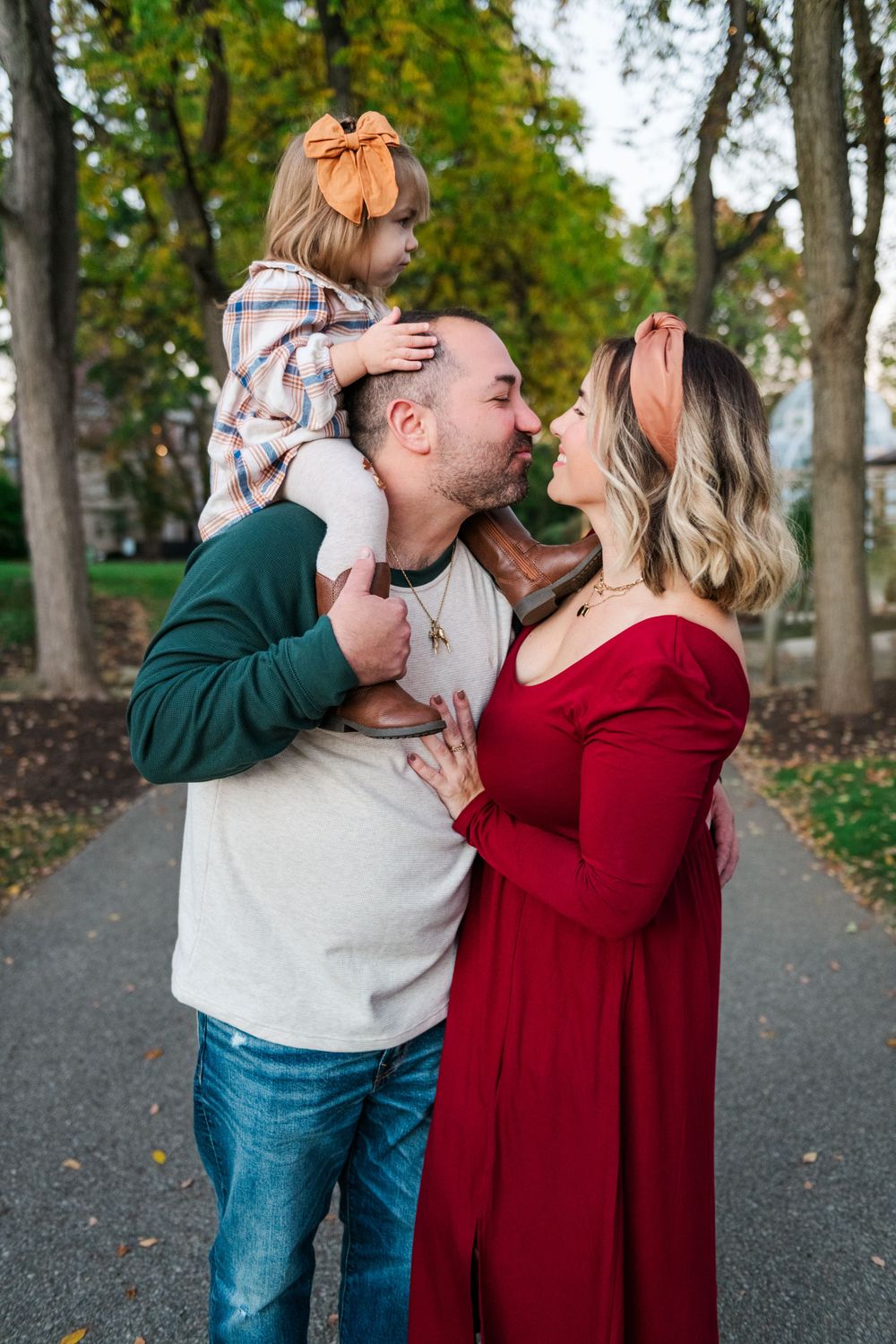 Intimate family moment captured outdoors on tree-lined path during fall with burgundy dress and green sweater.