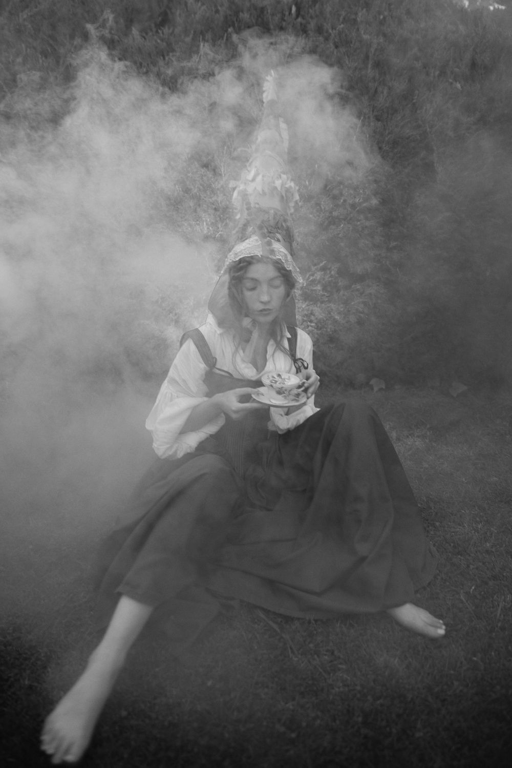 Black and white atmospheric photo of a figure in flowing dress sitting on grass surrounded by ethereal smoke in a moody scene.