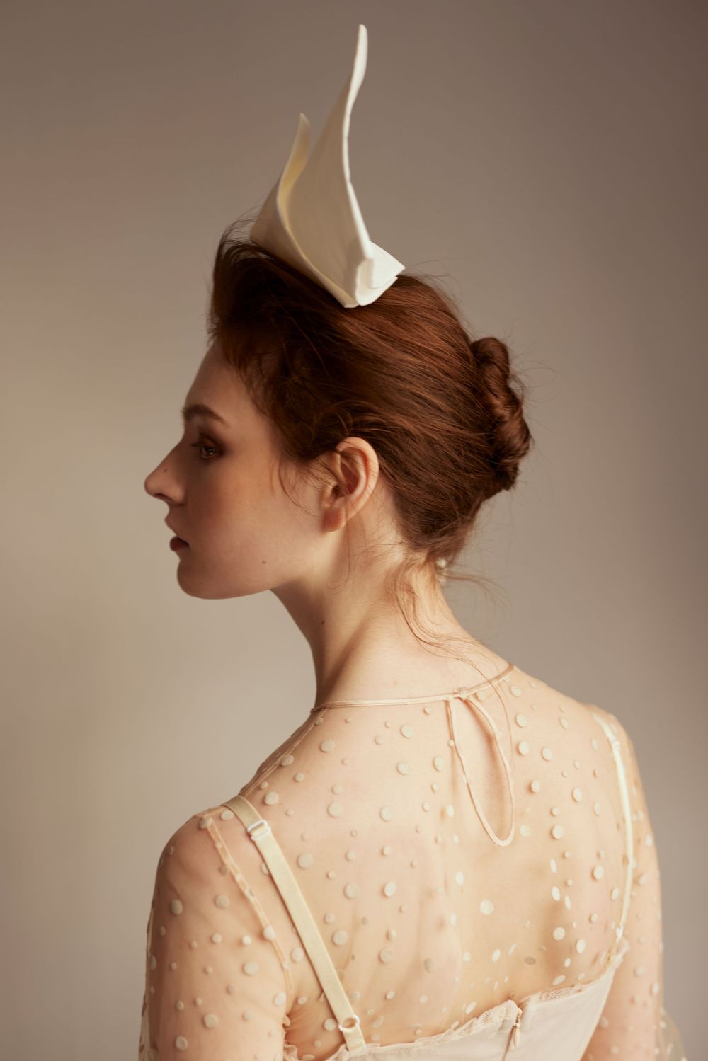 Profile view of a model in vintage-style cream dress with sheer dotted overlay and sculptural white headpiece against neutral backdrop.