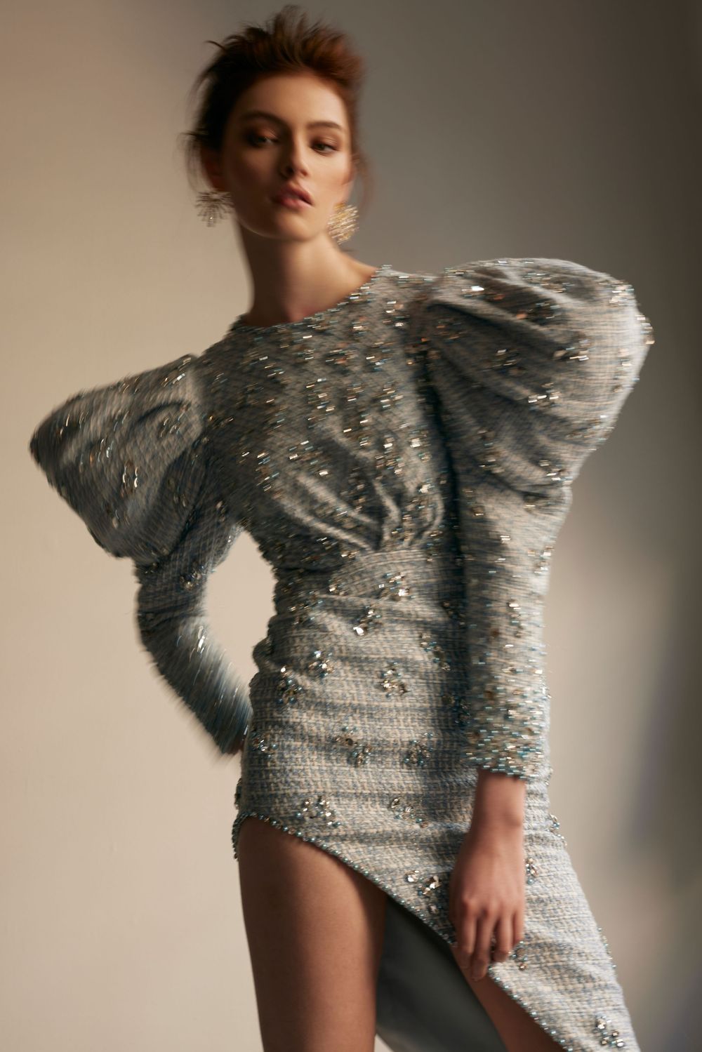 Fashion model wearing a dramatic silver sequined dress with exaggerated puff sleeve in artistic studio lighting.