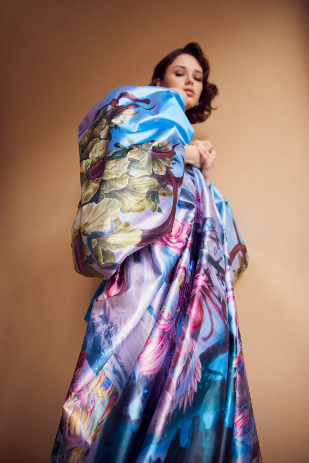 Figure draped in flowing blue and pink silk fabric with artistic floral pattern against bronze background.
