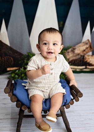 Baby in cream-colored romper sits happily on blue cushioned wooden bench with mountain-themed backdrop setup. #Milestone #6monthPictures #NM