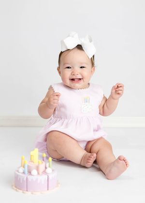 A baby in a pink dress with a white bow sits beside a decorative pink birthday cake on a white background. #CarlsbadNM #FirstBirthdayPhoto