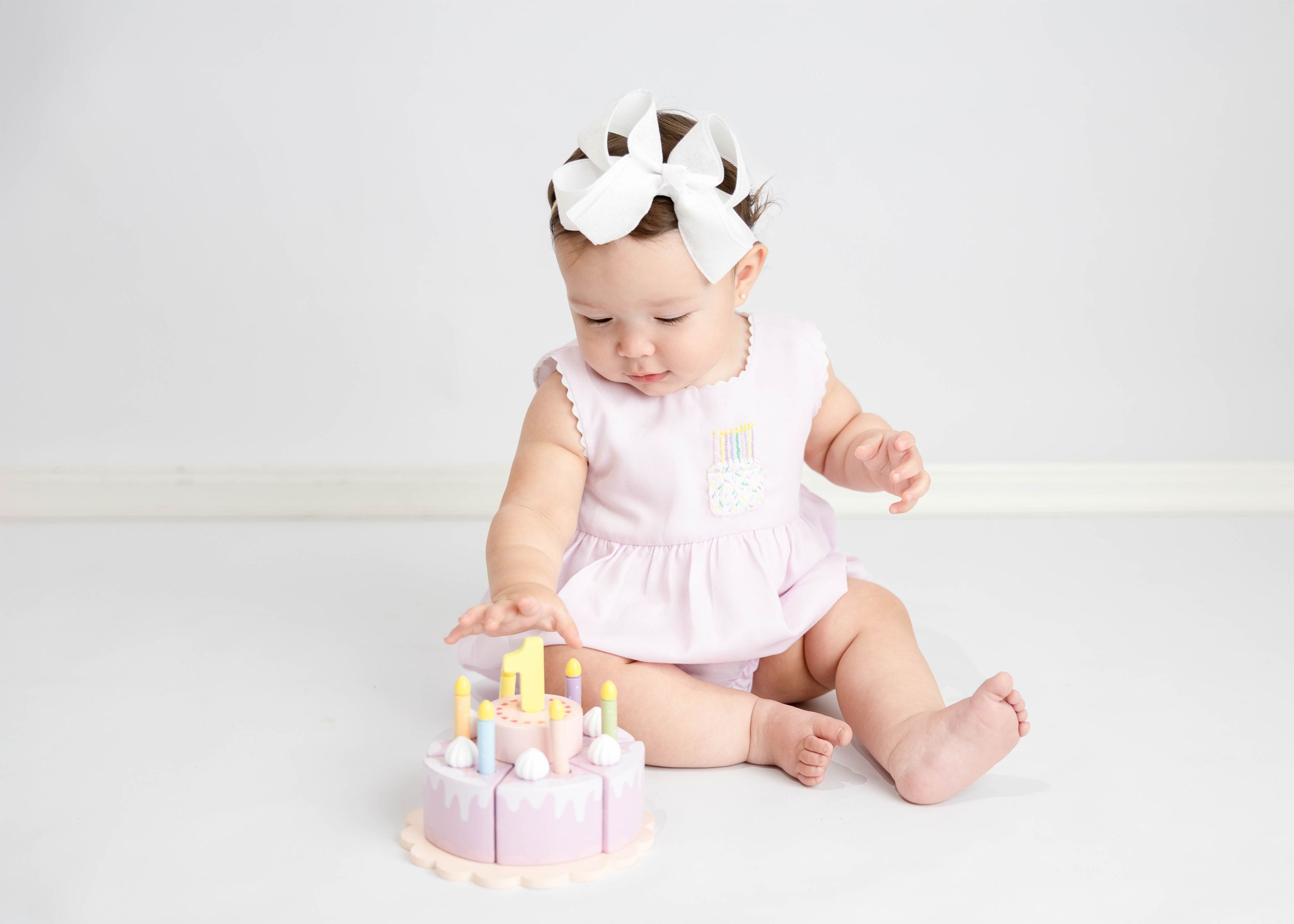 Small baby in pink dress with bow sits on white floor playing with pink and yellow wooden toy birthday cake. #FirstBirthday #CarlsbadNM #NM