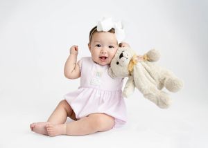 A baby in a pink dress sits on a white background while holding a plush teddy bear toy. #Carlsbad #NewMexico #childphotographer #babypicture