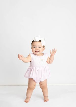 A baby in a pink romper and bow stands with arms raised looking happy against a white background. #firstyearpictures #Carlsbad #Artesia #NM