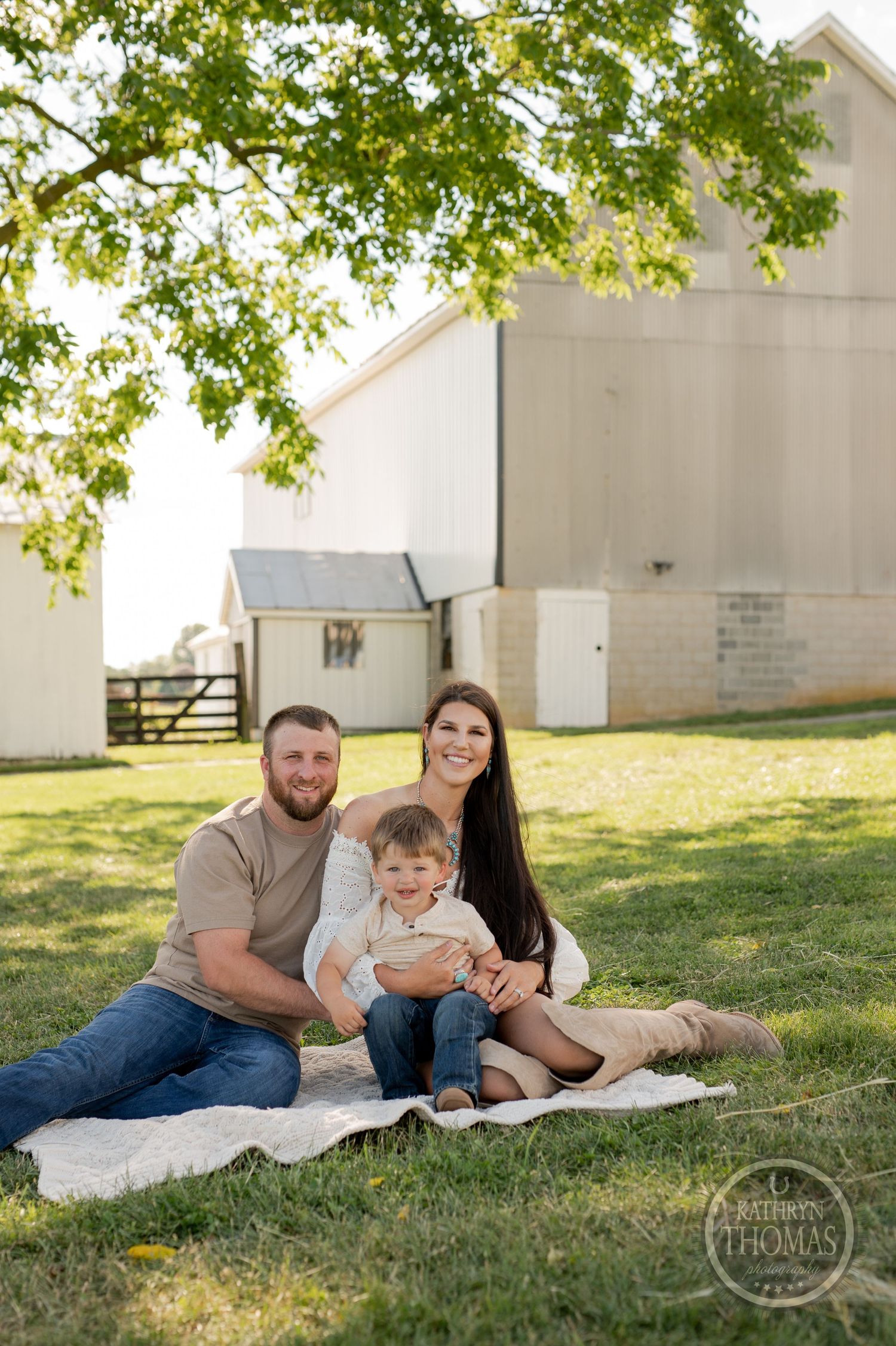 Family posing on blanket outdoors with white barn and green trees in background.