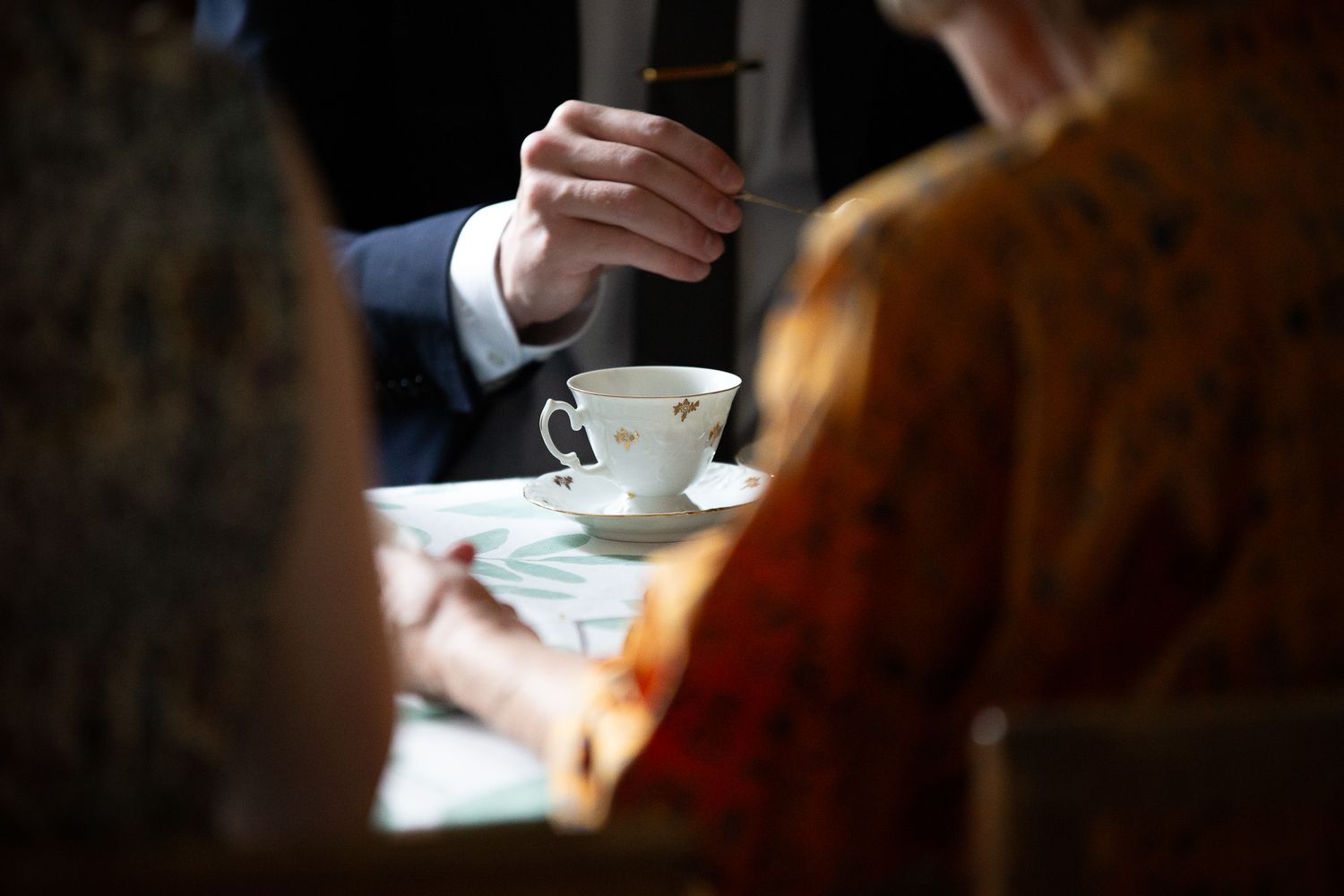 Lähikuva kahdesta kädestä pöydän ääressä, jonka keskellä on teekuppi aluslautasineen. Toinen käsi pitelee pientä lusikkaa.