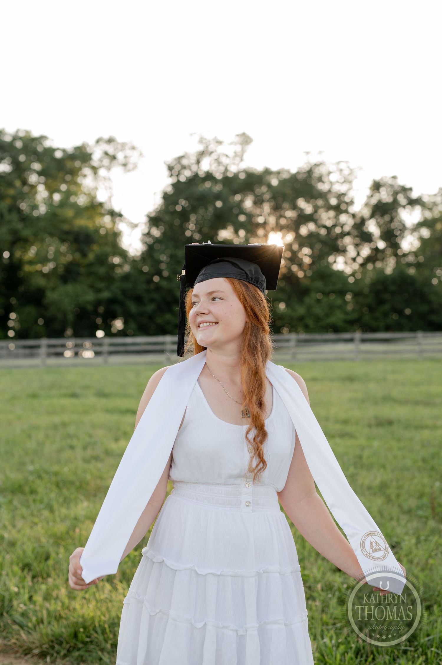 Graduate in white dress and cap smiles joyfully in sunlit field with white fence.