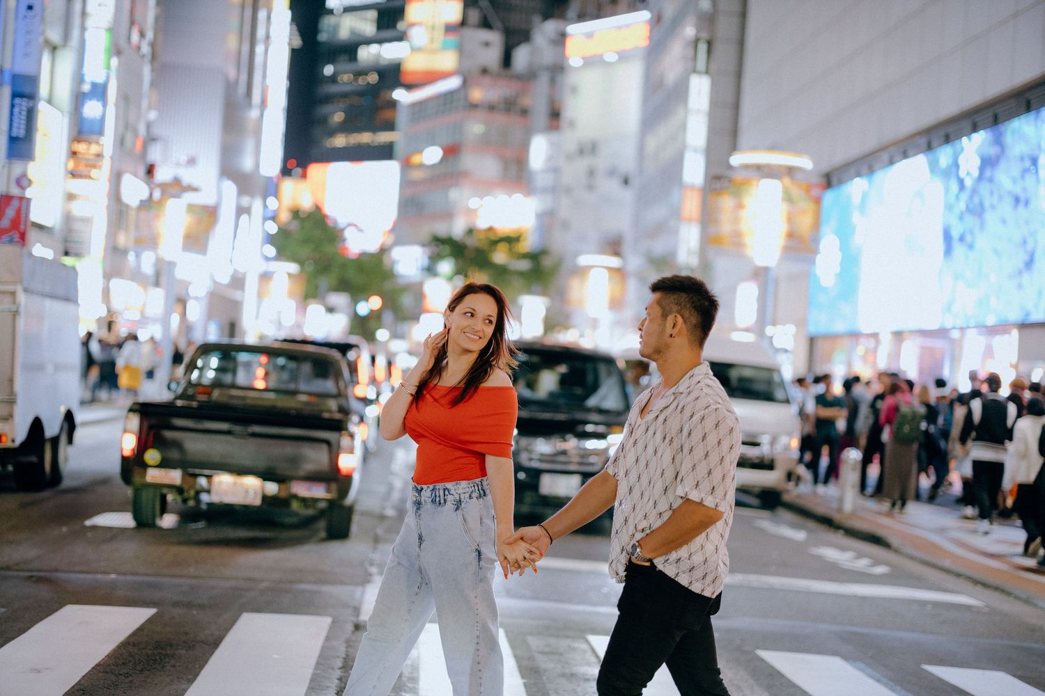 Two people walking on a busy city street at night with illuminated buildings and billboards in Tokyo.