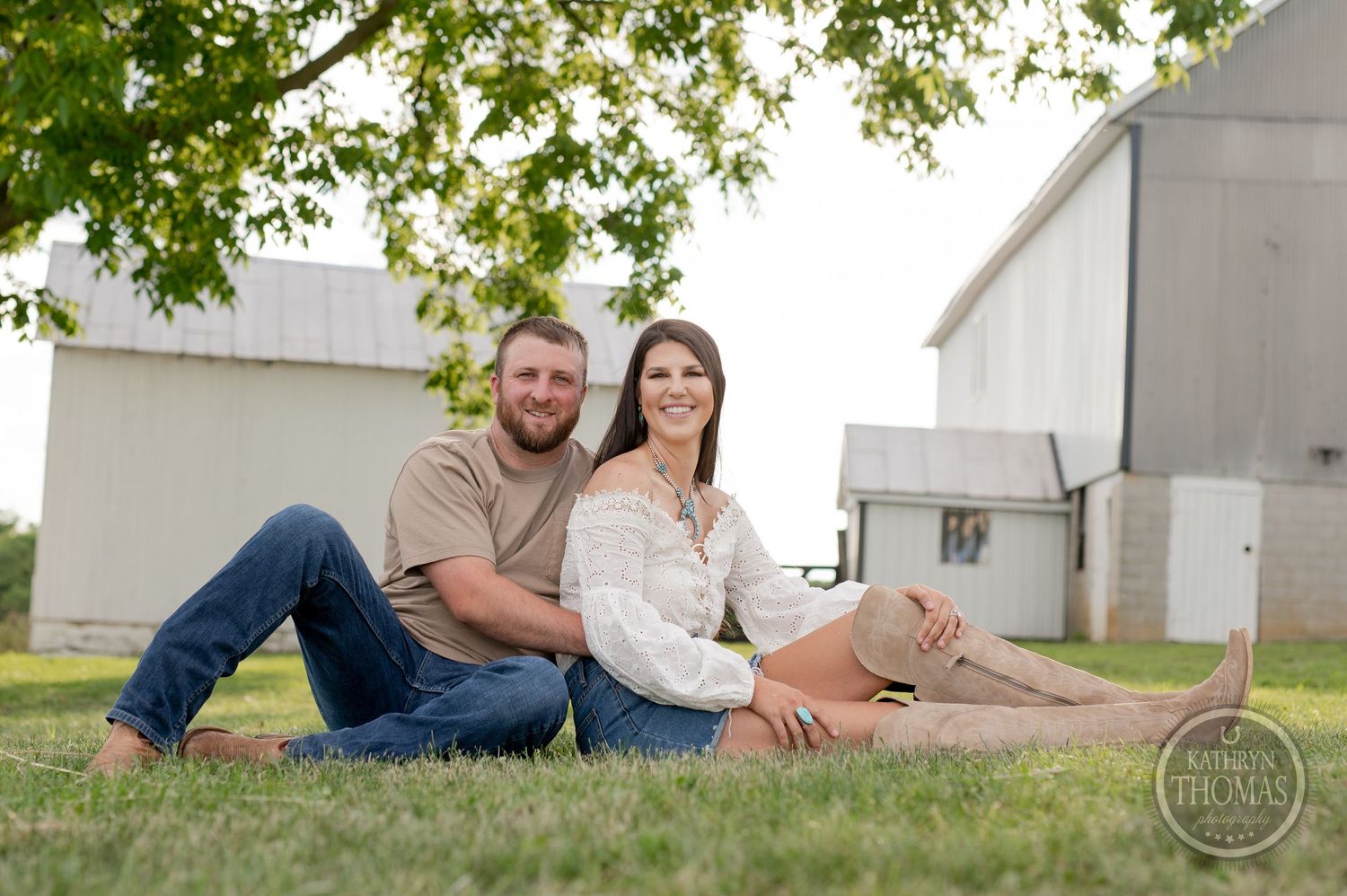 A couple relaxes together on a grassy lawn with farm buildings in the background during a sunny day.