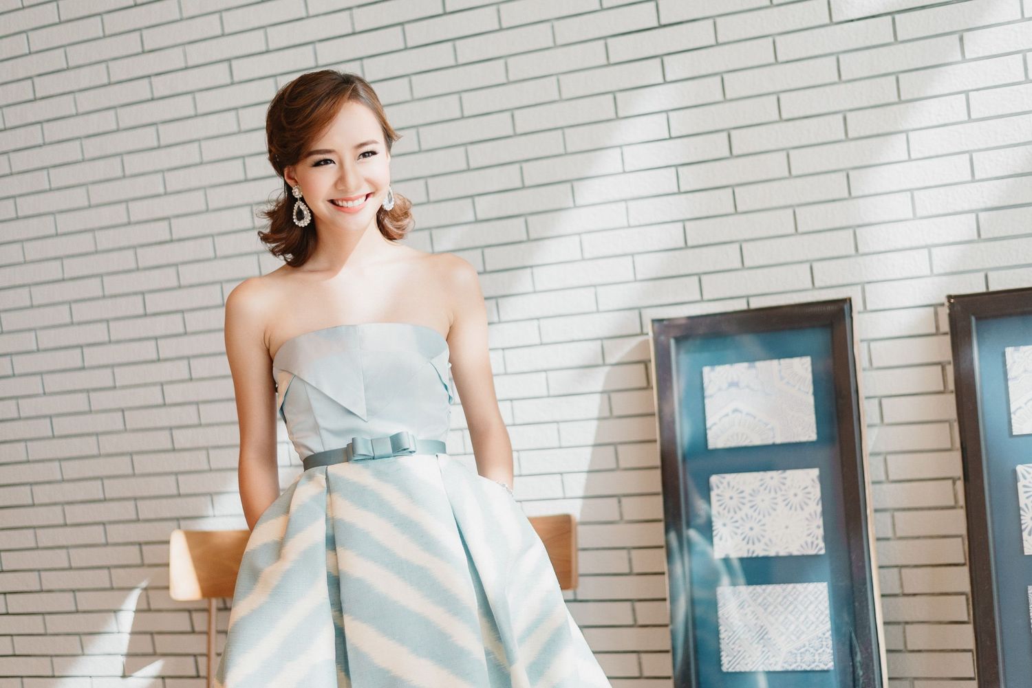 Elegant woman in strapless striped dress poses against white brick wall.