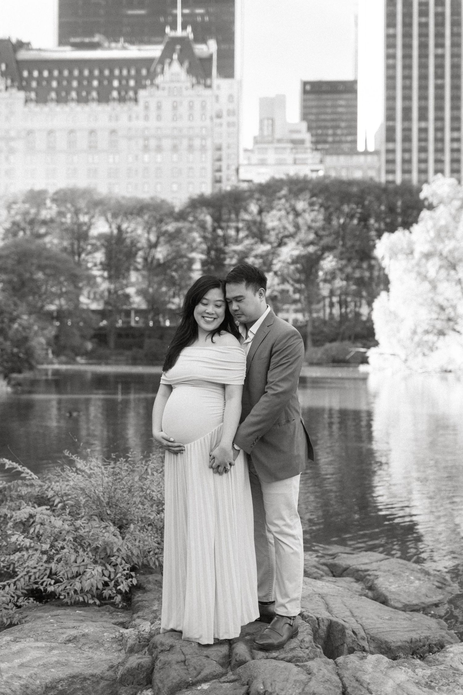 Couple poses for romantic photos by a lake with city skyline in background.