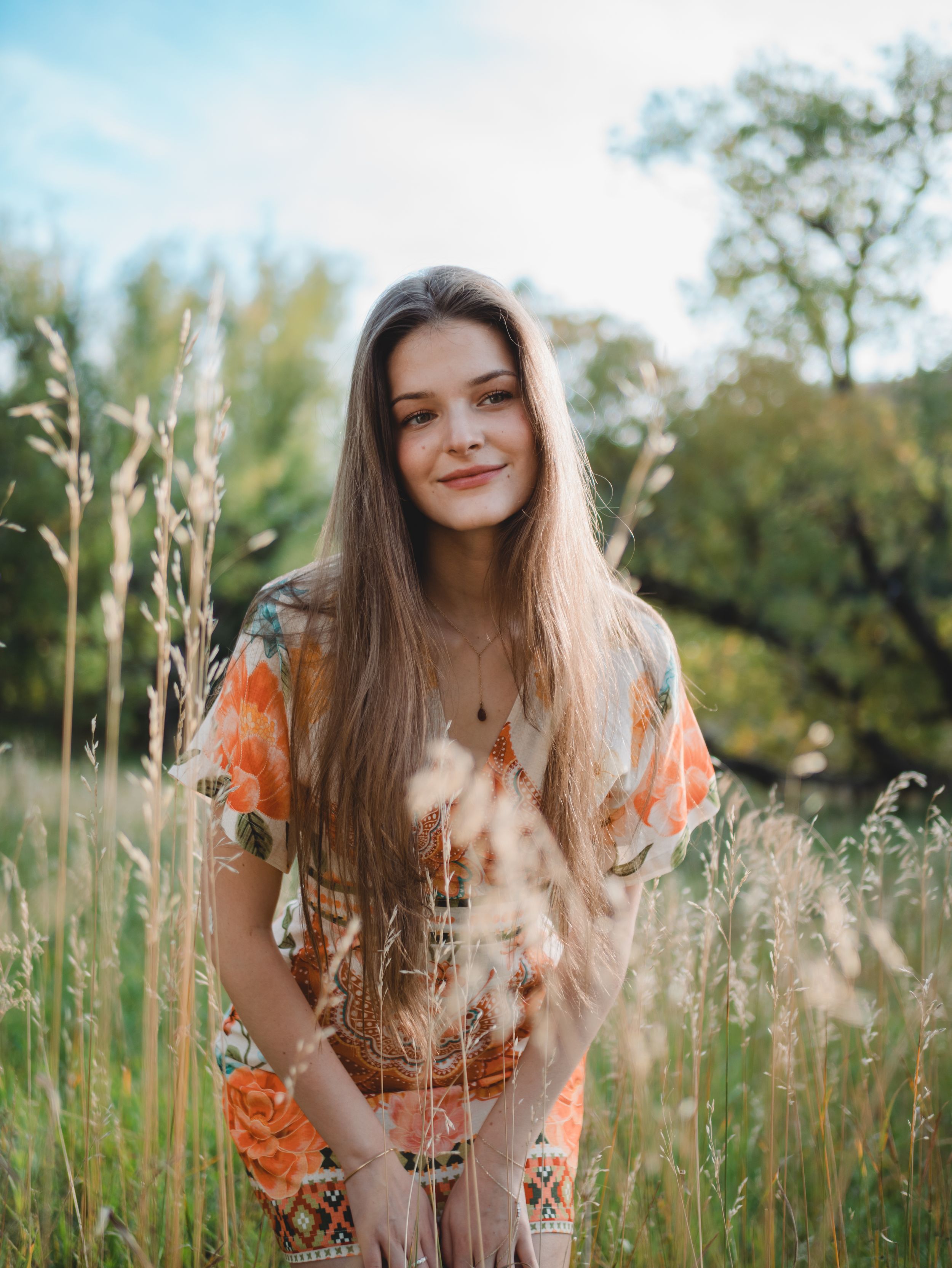 A person in an orange floral dress stands in a summer meadow with tall grass and trees in the background.