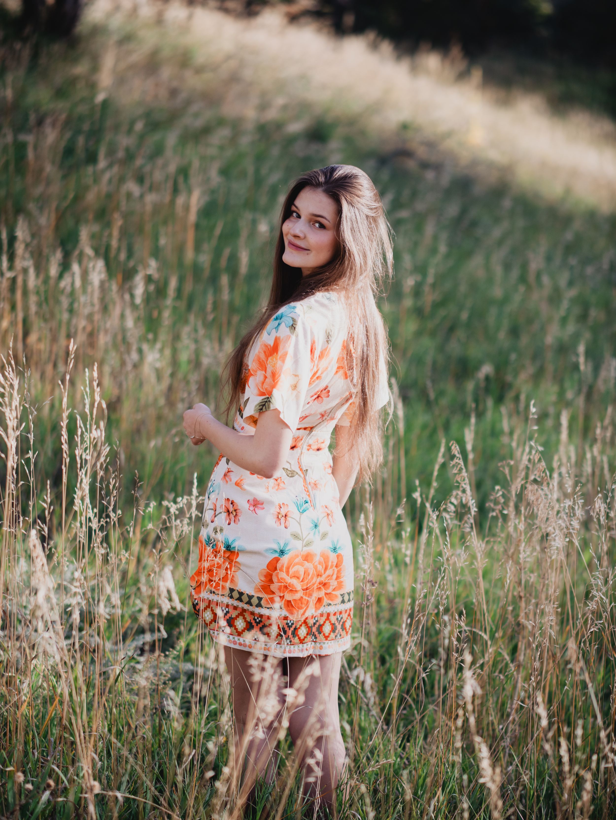 A person in a floral top poses gracefully in a sunlit meadow of tall grass during golden hour.