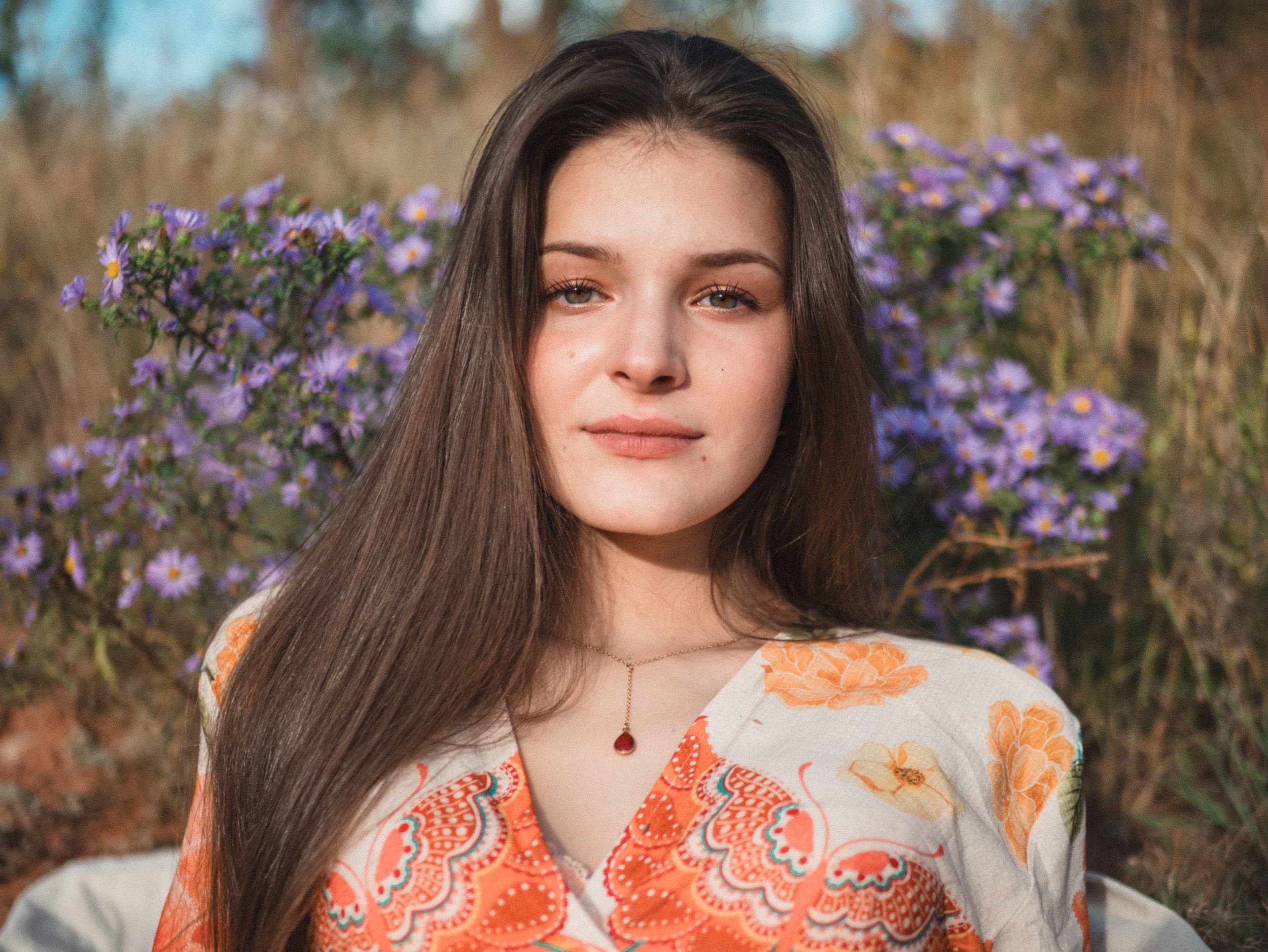 A woman in an orange and white patterned dress poses in a field of purple wildflowers on a sunny day.