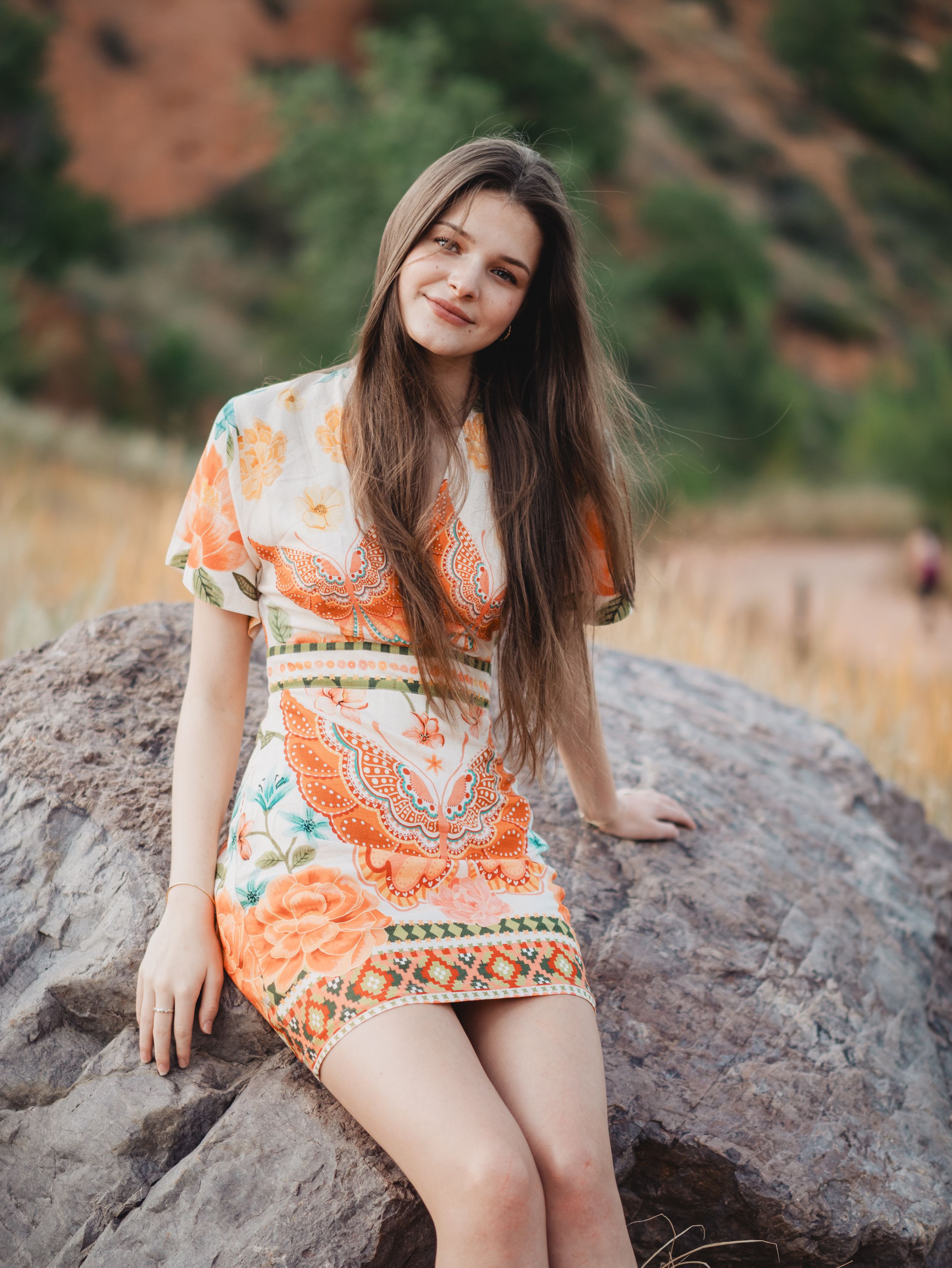 A woman in an orange and white patterned dress sits on red rocks in a natural outdoor setting.