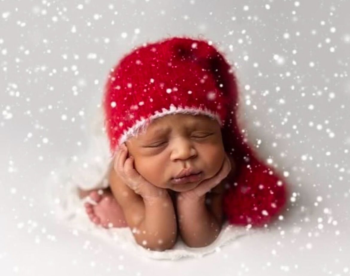 Sleeping baby wearing red knit hat on white blanket with falling snow effect in studio portrait.